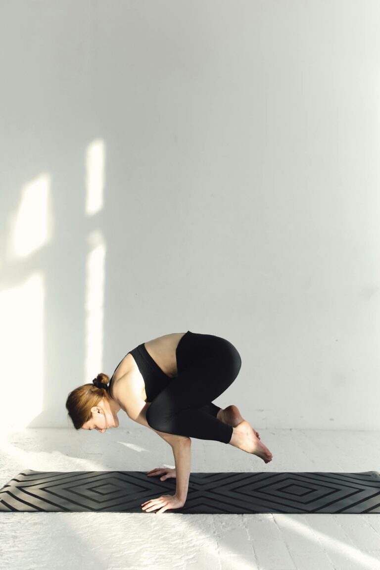 Woman in black activewear practicing crow pose on a yoga mat indoors, emphasizing balance and mindfulness.